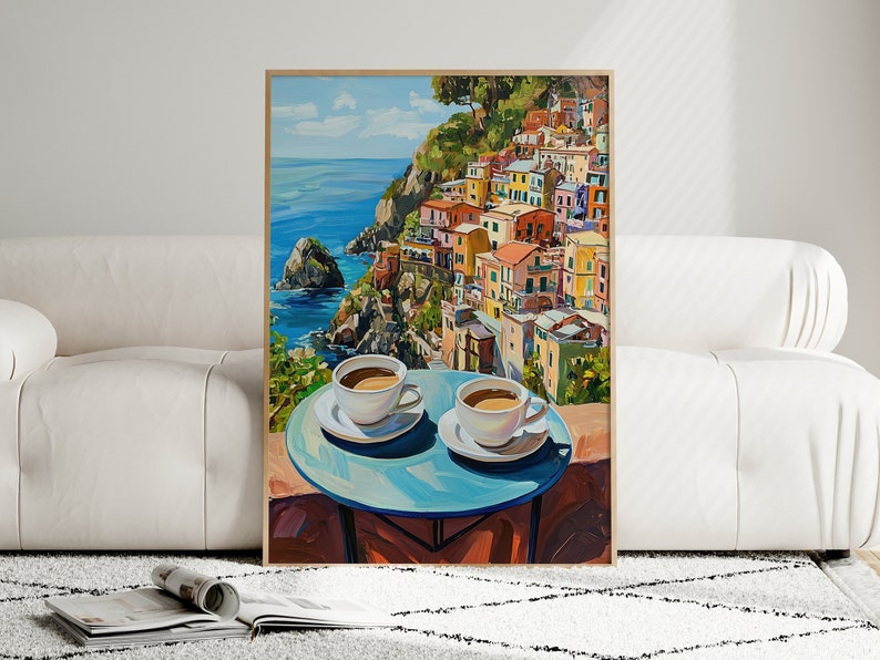 May include: A painting of a colorful Italian village on a cliff overlooking the ocean. Two cups of coffee sit on a blue table in the foreground.