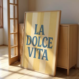 May include: A framed poster with the words "LA DOLCE VITA" in blue letters against a yellow and white striped background. The poster is leaning against a light wood cabinet in a room with a wooden floor and a window.
