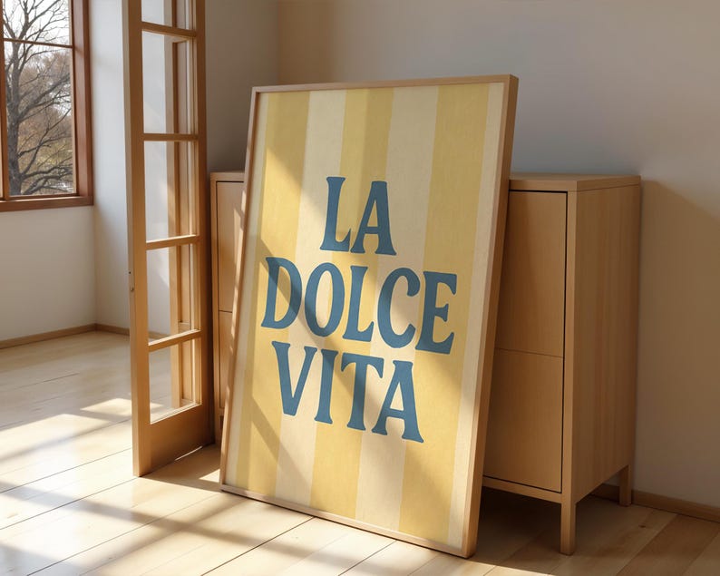 May include: A framed poster with the words "LA DOLCE VITA" in blue letters against a yellow and white striped background. The poster is leaning against a light wood cabinet in a room with a wooden floor and a window.