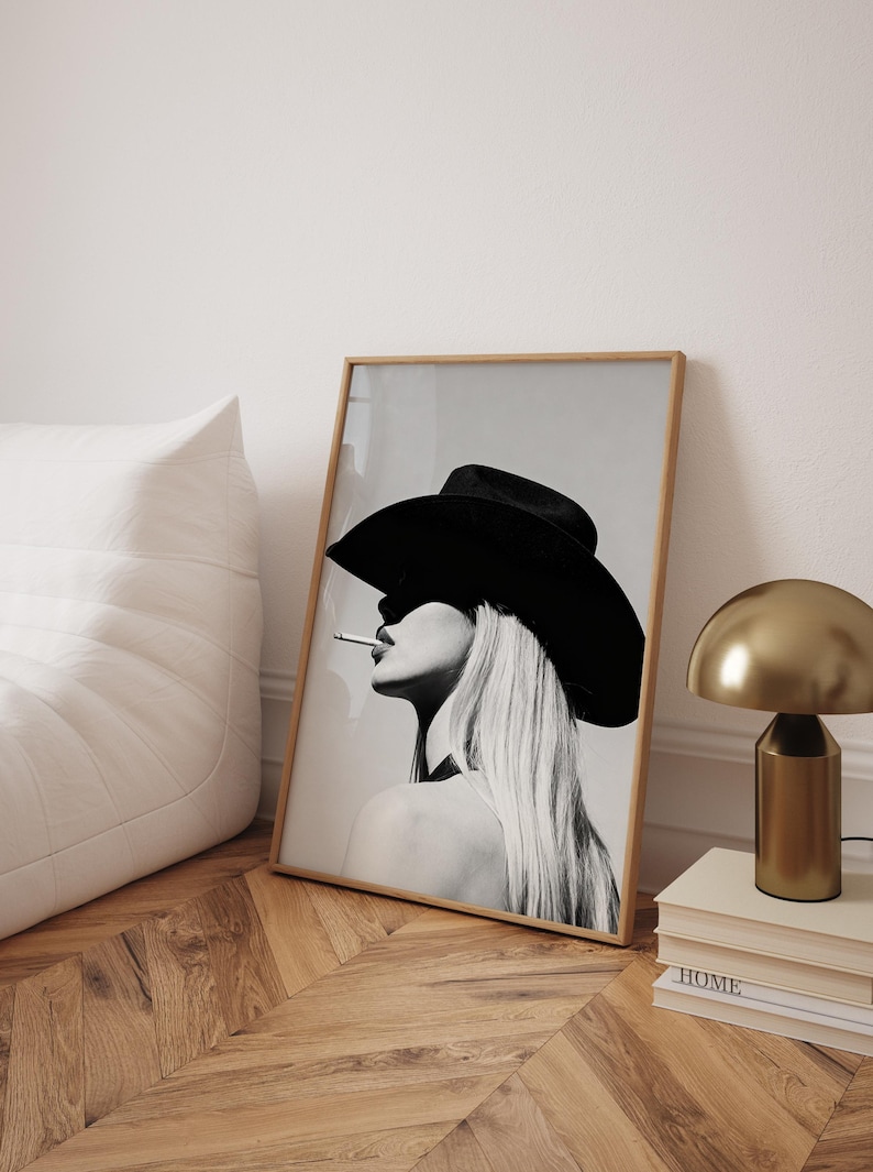 May include: Black and white framed art print featuring a woman wearing a large black cowboy hat and smoking a cigarette. The artwork has a minimalist aesthetic, with long blonde hair and a neutral background. The print is displayed on a wooden floor.