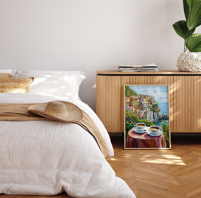 May include: A painting of a colorful Italian village on a cliff overlooking the ocean. Two cups of coffee are on a table in the foreground.