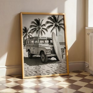 May include: Black and white framed print featuring a vintage SUV parked on a sandy beach with a surfboard leaning against it. Palm trees are in the background, creating a coastal theme. The print is in a wooden frame.