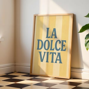 May include: A framed art print with the words "LA DOLCE VITA" in blue lettering on a yellow and cream striped background. The print is in a light wood frame and leans against a wall. A potted plant is to the right.