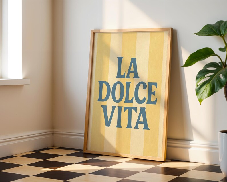May include: A framed art print with the words "LA DOLCE VITA" in blue lettering on a yellow and cream striped background. The print is in a light wood frame and leans against a wall. A potted plant is to the right.