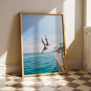 May include: A framed print depicting two people leaping into turquoise water from a boat. The wooden frame surrounds the image of a sunny day with a blue sky. The boat's deck is visible on the right side of the image.
