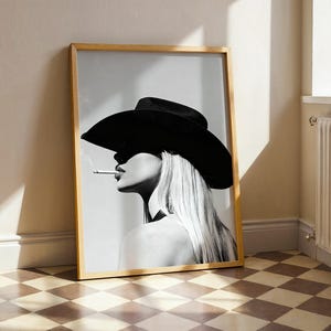 May include: Black and white framed print featuring a woman wearing a black cowboy hat and smoking a cigarette. The artwork is leaning against a wall, with a light wood frame and a checkered floor.
