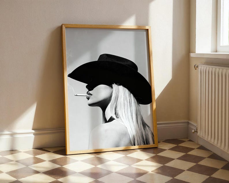 May include: Black and white framed print featuring a woman wearing a black cowboy hat and smoking a cigarette. The artwork is leaning against a wall, with a light wood frame and a checkered floor.