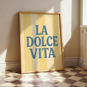 May include: A framed art print with the words "LA DOLCE VITA" in blue lettering on a yellow and white striped background. The frame is a light brown color, and the artwork is leaning against a white wall. The floor has a checkered pattern.