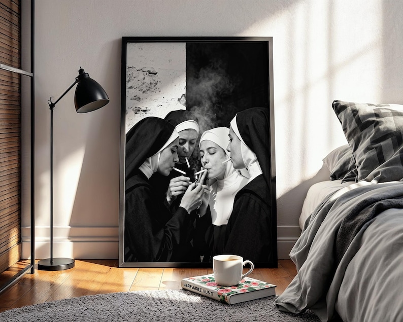 May include: Black and white framed print featuring four nuns in traditional habits, smoking cigarettes. The image is set against a black and white backdrop, with smoke rising. A coffee cup and a book are in the foreground, resting on a gray rug.