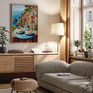 May include: A framed painting of a colorful Italian village on a cliff overlooking the ocean. Two cups of coffee are on a table in the foreground. The painting is hanging on a wall in a living room with a couch, a side table, and plants.