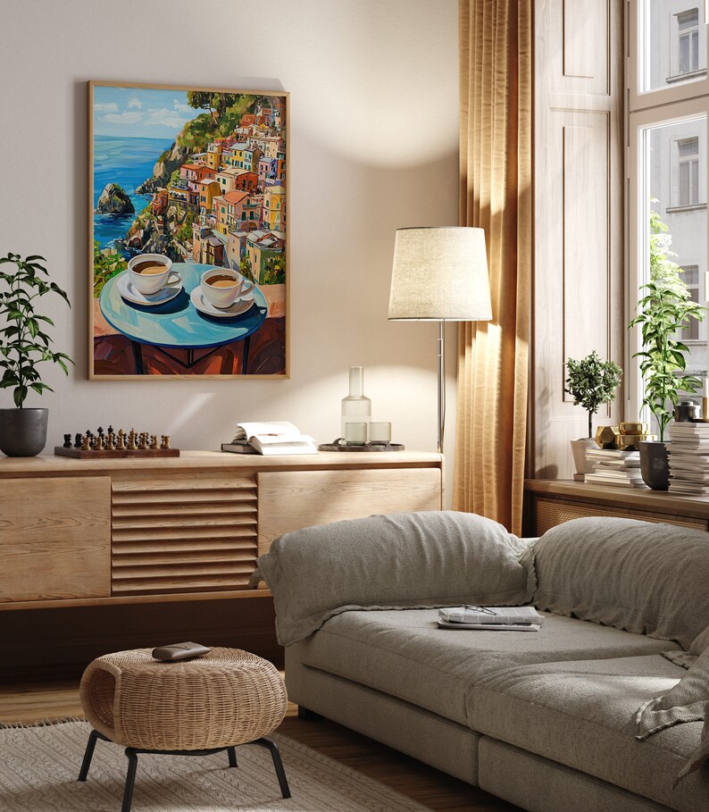 May include: A framed painting of a colorful Italian village on a cliff overlooking the ocean. Two cups of coffee are on a table in the foreground. The painting is hanging on a wall in a living room with a couch, a side table, and plants.