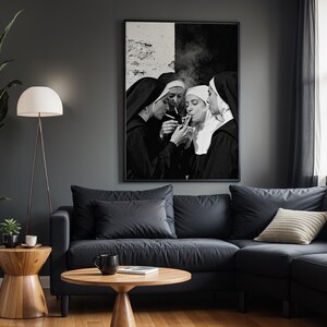 May include: Black and white framed art print depicting four nuns smoking cigarettes. The artwork hangs above a dark gray sectional sofa with a light beige pillow. A wooden side table and floor lamp are in the foreground.