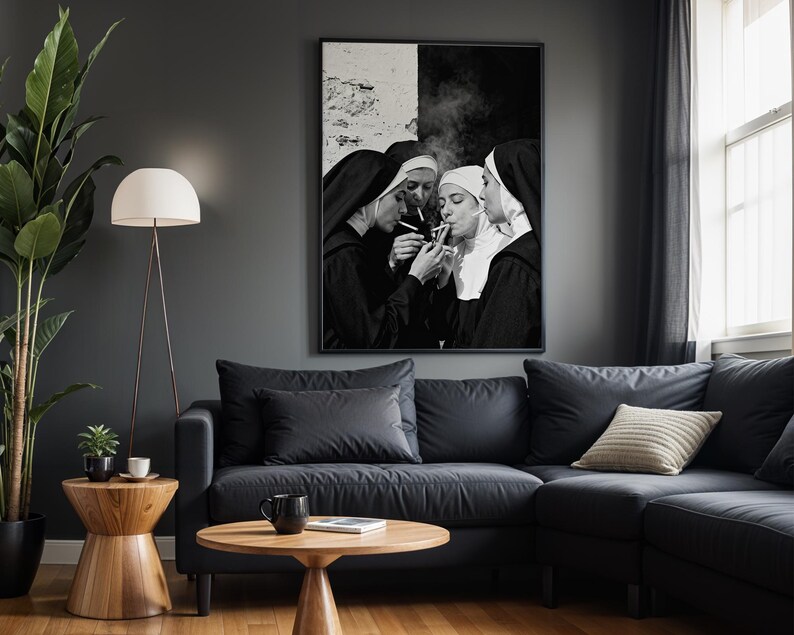 May include: Black and white framed art print depicting four nuns smoking cigarettes. The artwork hangs above a dark gray sectional sofa with a light beige pillow. A wooden side table and floor lamp are in the foreground.