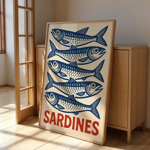 Może przedstawiać: Oprawiony wydruk z sześcioma stylizowanymi niebieskimi rybami na kremowym tle. Słowo "SARDINES" jest wydrukowane na czerwono u dołu. Grafika opiera się o jasną drewnianą szafkę.