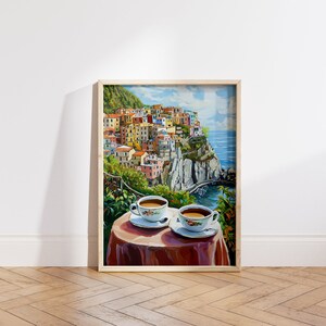 May include: A framed painting of a colorful Italian village perched on a cliff overlooking the ocean. Two white coffee cups with brown coffee sit on a red tablecloth in the foreground.