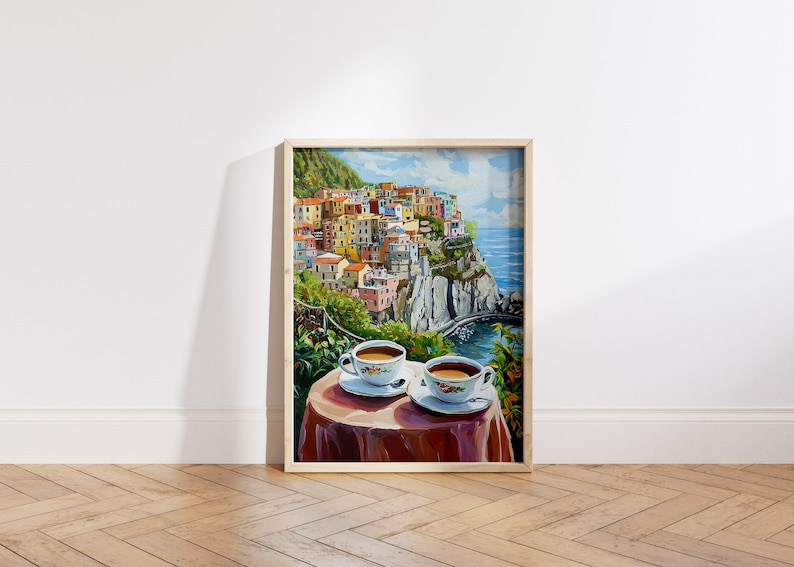 May include: A framed painting of a colorful Italian village perched on a cliff overlooking the ocean. Two white coffee cups with brown coffee sit on a red tablecloth in the foreground.