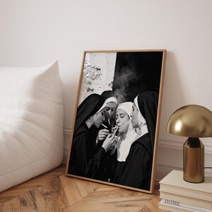 May include: Black and white framed print featuring four nuns in traditional habits, smoking cigarettes. The image is set against a textured wall and a dark background. The print is displayed on a wooden floor, next to a white beanbag chair and a gold lamp.