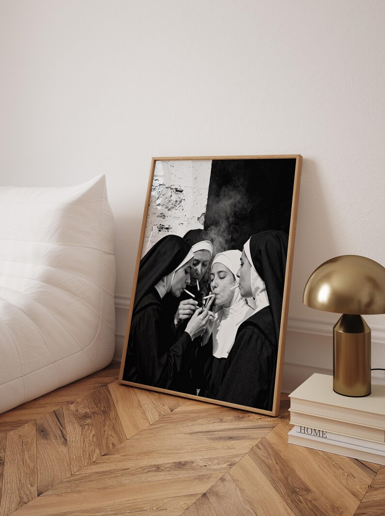 May include: Black and white framed print featuring four nuns in traditional habits, smoking cigarettes. The image is set against a textured wall and a dark background. The print is displayed on a wooden floor, next to a white beanbag chair and a gold lamp.