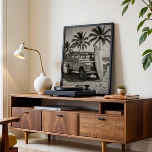May include: A mid-century modern style wooden media console with three drawers and a shelf. A framed black and white photograph of a vintage vehicle and surfboard is on top. A white lamp and a record player are also on the console.
