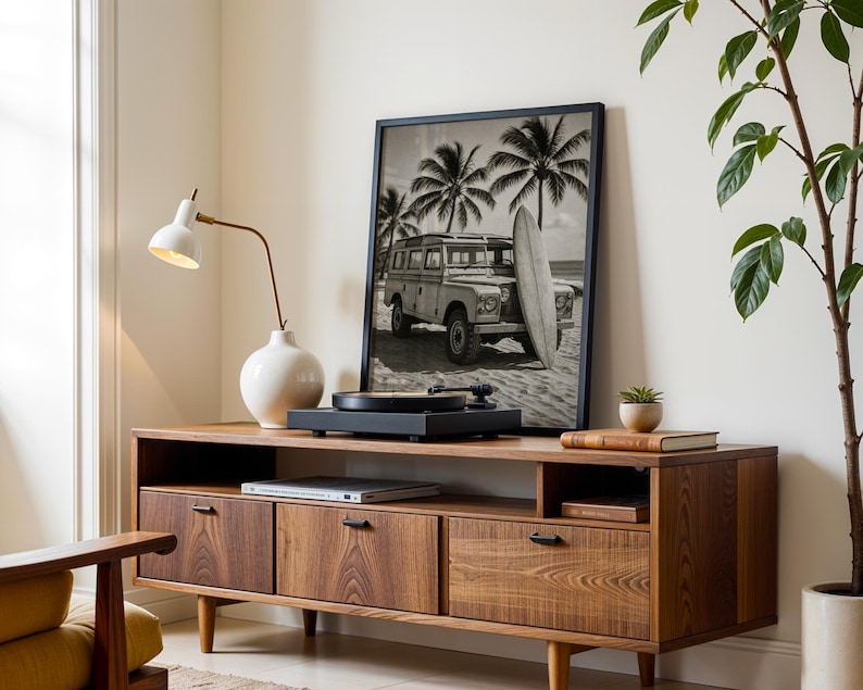 May include: A mid-century modern style wooden media console with three drawers and a shelf. A framed black and white photograph of a vintage vehicle and surfboard is on top. A white lamp and a record player are also on the console.
