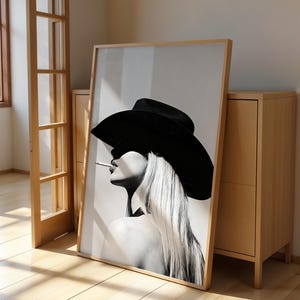 May include: Black and white framed art print featuring a woman in a black hat with long blonde hair, smoking a cigarette. The artwork is leaning against a light wood cabinet and a wooden door frame.