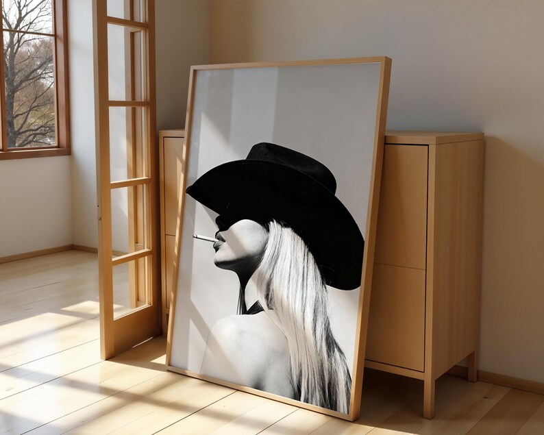 May include: Black and white framed art print featuring a woman in a black hat with long blonde hair, smoking a cigarette. The artwork is leaning against a light wood cabinet and a wooden door frame.