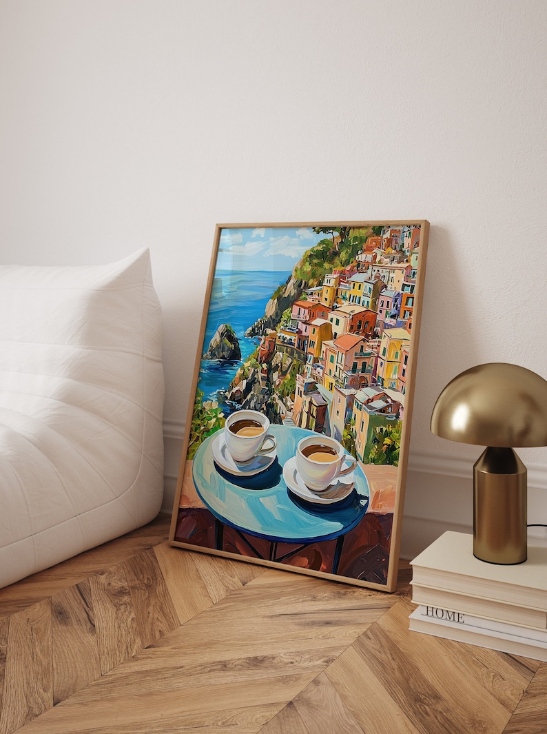 May include: A framed painting of a colorful Italian village overlooking the ocean. Two white coffee cups with saucers are on a blue table in the foreground.
