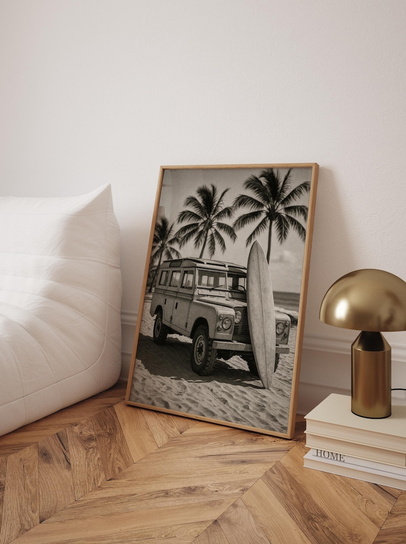 May include: Black and white framed print of a vintage SUV parked on a sandy beach with a surfboard leaning against it. Palm trees are in the background, evoking a tropical, coastal theme.