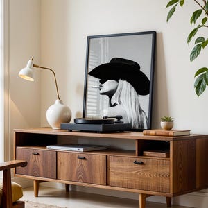 May include: A mid-century modern style wooden media console with three drawers and a shelf. A framed black and white portrait of a woman wearing a hat is on top, along with a record player and a white lamp.