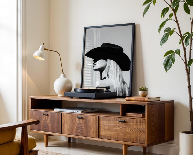May include: A mid-century modern style wooden media console with three drawers and a shelf. A framed black and white portrait of a woman wearing a hat is on top, along with a record player and a white lamp.