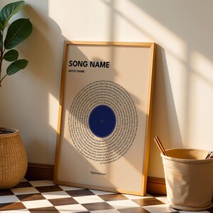 May include: A framed print with a beige background features a circular design of song lyrics around a blue circle. The text "SONG NAME" and "ARTIST NAME" are at the top. A woven basket with a plant and a beige bucket are also in the image.