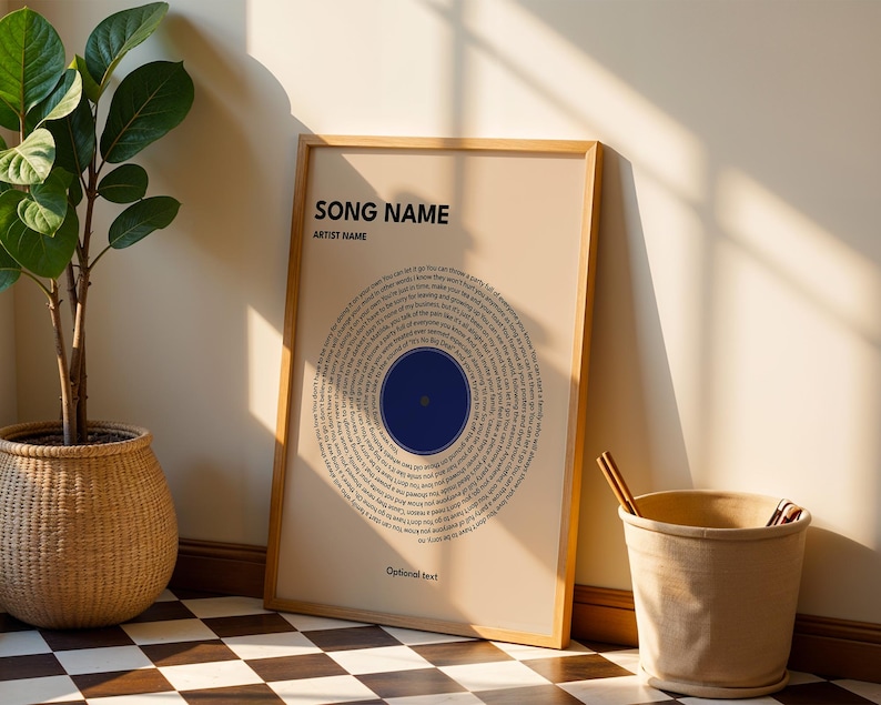 May include: A framed print with a beige background features a circular design of song lyrics around a blue circle. The text "SONG NAME" and "ARTIST NAME" are at the top. A woven basket with a plant and a beige bucket are also in the image.
