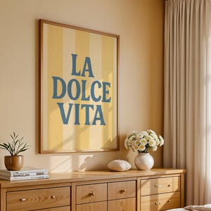 May include: A framed art print with the words "LA DOLCE VITA" in blue lettering on a yellow and white striped background. The artwork is displayed above a wooden dresser with a plant, books, and a vase of flowers.