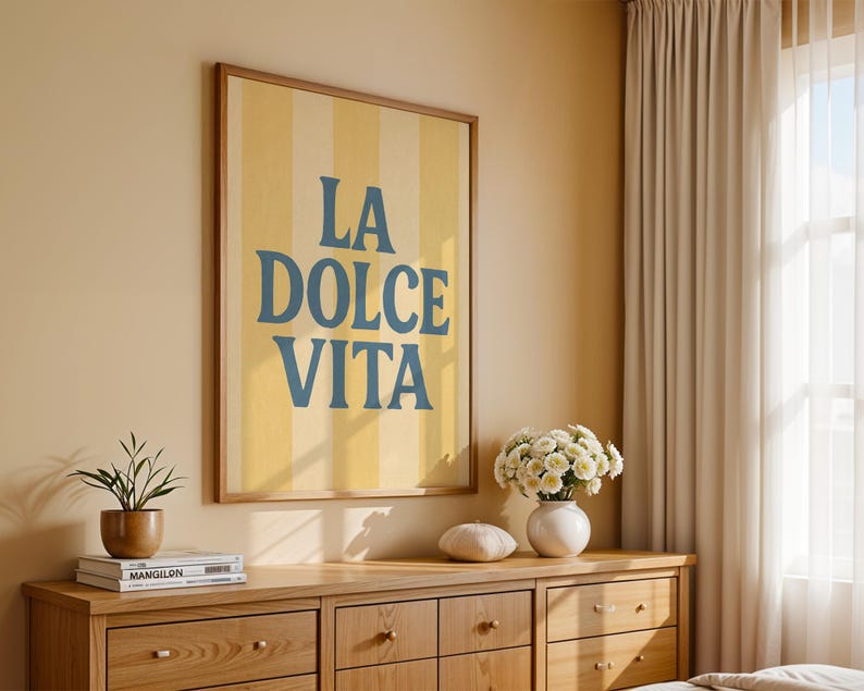 May include: A framed art print with the words "LA DOLCE VITA" in blue lettering on a yellow and white striped background. The artwork is displayed above a wooden dresser with a plant, books, and a vase of flowers.
