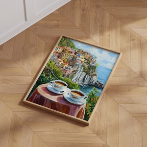 May include: A framed painting of a colorful Italian village perched on a cliff overlooking the ocean. Two white coffee cups with saucers are on a table in the foreground, with a red tablecloth and green foliage.