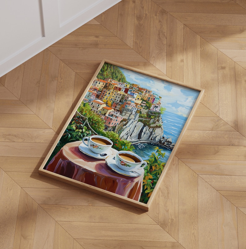 May include: A framed painting of a colorful Italian village perched on a cliff overlooking the ocean. Two white coffee cups with saucers are on a table in the foreground, with a red tablecloth and green foliage.