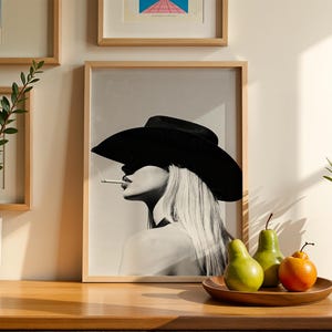 May include: Black and white framed print of a woman wearing a large black hat and smoking a cigarette. The artwork is displayed on a wooden surface with a wooden tray holding green pears and an orange fruit. Decorative plants are in the background.