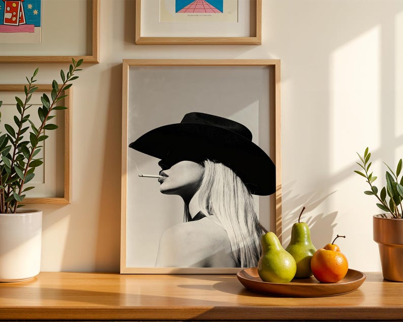 May include: Black and white framed print of a woman wearing a large black hat and smoking a cigarette. The artwork is displayed on a wooden surface with a wooden tray holding green pears and an orange fruit. Decorative plants are in the background.