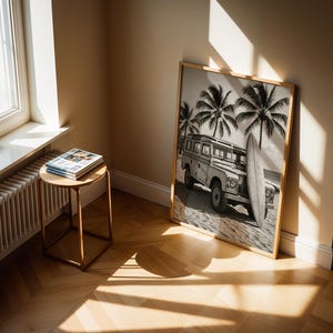 May include: A framed black and white print of a vintage SUV parked on a beach with a surfboard and palm trees. The print is leaning against a wall. A small round table with magazines sits near a window.