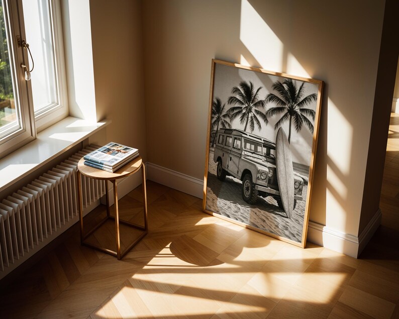 May include: A framed black and white print of a vintage SUV parked on a beach with a surfboard and palm trees. The print is leaning against a wall. A small round table with magazines sits near a window.