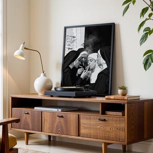 May include: A mid-century modern style wooden media console with a framed black and white print of nuns smoking. A white lamp and a white vase are on the console. A record player sits on top of the console.
