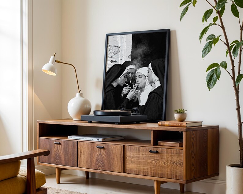May include: A mid-century modern style wooden media console with a framed black and white print of nuns smoking. A white lamp and a white vase are on the console. A record player sits on top of the console.