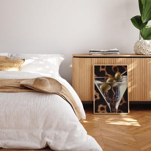 May include: A white bed with a tan blanket and a brown hat on top. A wooden cabinet with a framed print of a martini glass with olives on a leopard print background.