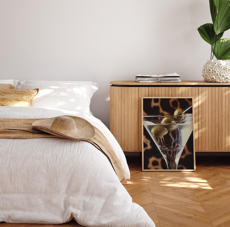 May include: A white bed with a tan blanket and a brown hat on top. A wooden cabinet with a framed print of a martini glass with olives on a leopard print background.