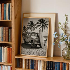 May include: Black and white framed print featuring a vintage SUV parked on a sandy beach with a surfboard and palm trees. The print is displayed on a wooden shelf next to a bookshelf filled with books and a vase of blue flowers.