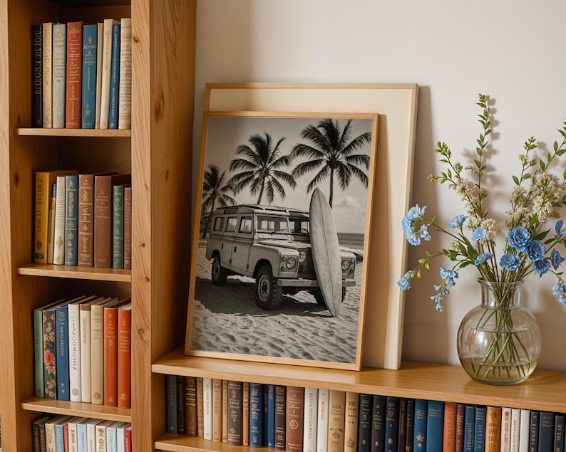 May include: Black and white framed print featuring a vintage SUV parked on a sandy beach with a surfboard and palm trees. The print is displayed on a wooden shelf next to a bookshelf filled with books and a vase of blue flowers.