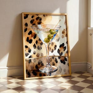 Puede incluir: Impresión enmarcada que presenta una ilustración de acuarela de un martini con aceitunas. La copa de cóctel está centrada, con un fondo de estampado de leopardo en tonos marrón, negro y beige. El marco es de color madera clara.