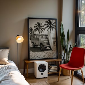 May include: A room with a black and white framed print of a vintage SUV on a beach with palm trees. A white speaker sits below the print on a wooden bench. A red chair and a floor lamp are also in the room.