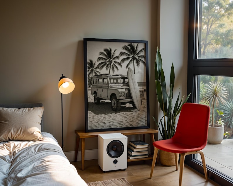 May include: A room with a black and white framed print of a vintage SUV on a beach with palm trees. A white speaker sits below the print on a wooden bench. A red chair and a floor lamp are also in the room.