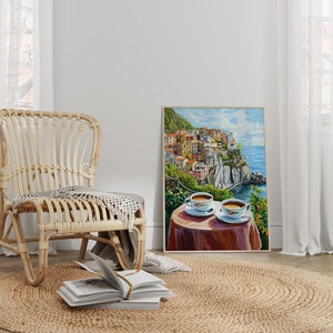 May include: A wicker chair with a throw blanket sits on a jute rug in front of a painting of a colorful Italian village on a cliff overlooking the ocean. Two cups of coffee are on a table in the painting.